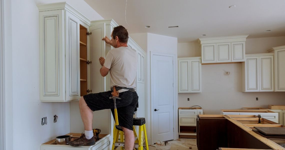 A carpenter standing on a small yellow stepladder to adjust and install a white upper cabinet door