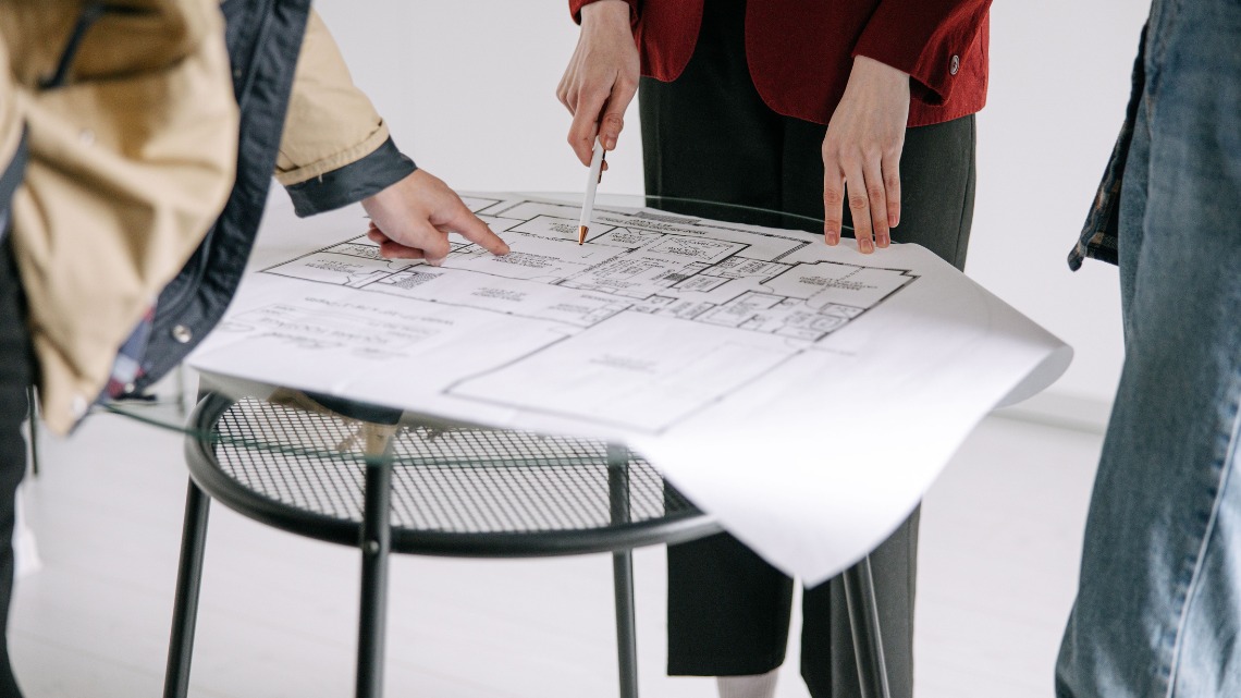 people stand around a glass table reviewing a printed architectural blueprint plan together